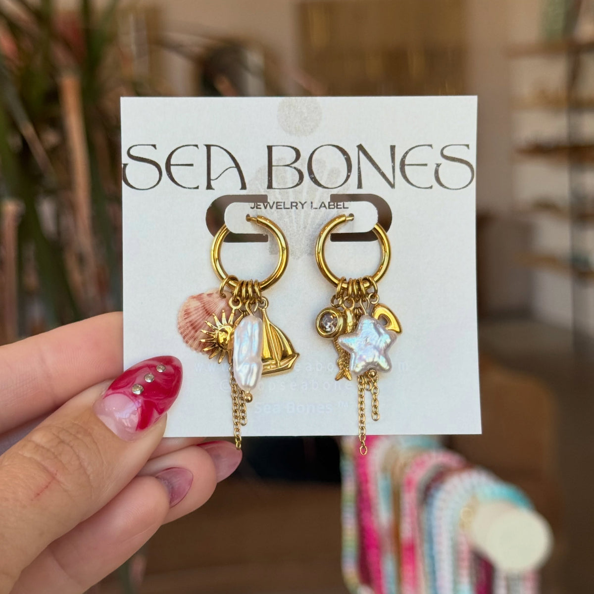 A hand with pink glitter nail polish holds a white display card featuring gold-tone earrings with shell, starfish, and pearl charms. The card is branded 'SEA BONES JEWELRY LABS'.